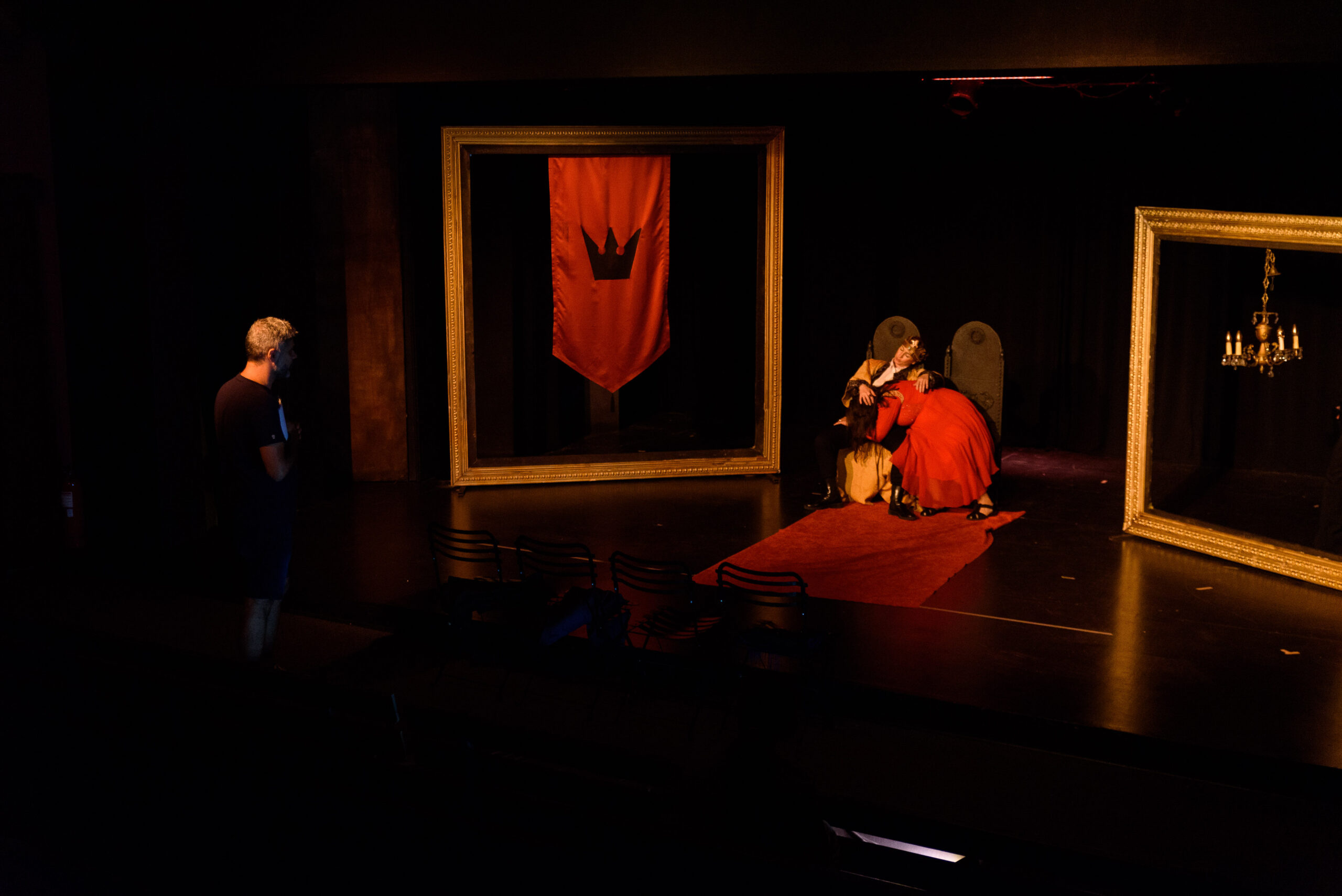 Σκηνή από Άμλετ Β', ένας βασιλιάς και μια βασίλισσα στο κέντρο της σκηνής, κάθονται στους θρόνους τους και ο ένας είναι στην αγκαλιά του άλλου. Πίσω τους δεξιά και αριστερά είναι δύο τεράστιες κορνίζες. Στο μέσα μέρος της αρστερής κρέμεται ένα λάβαρο με θυρεό, ενώ στη δεξιά ένας πολυέλαιος. Μπροστά και αριστερά στέκεται όρθιος ο σκηνοθέτης της παράστασης και παρακολουθεί