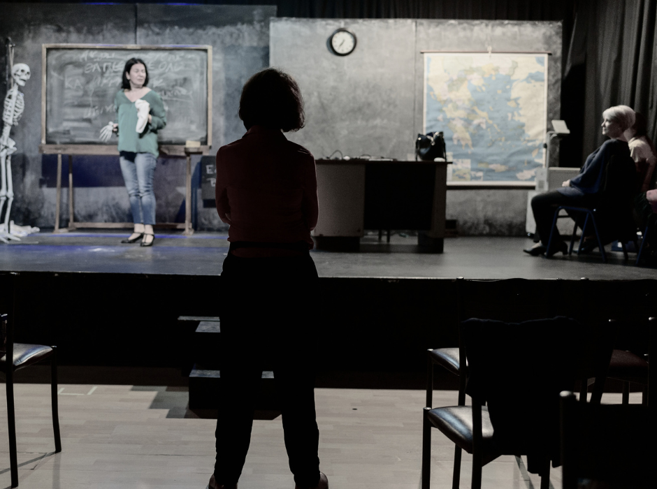 Μία γυναίκα σκηνοθέτης στέκεται στην πλατεία του θεάτρου, πλάτη στον φακό και κοιτάζει τη σκηνή όπου δύο ηθοποιοί κάνουν πρόβα.