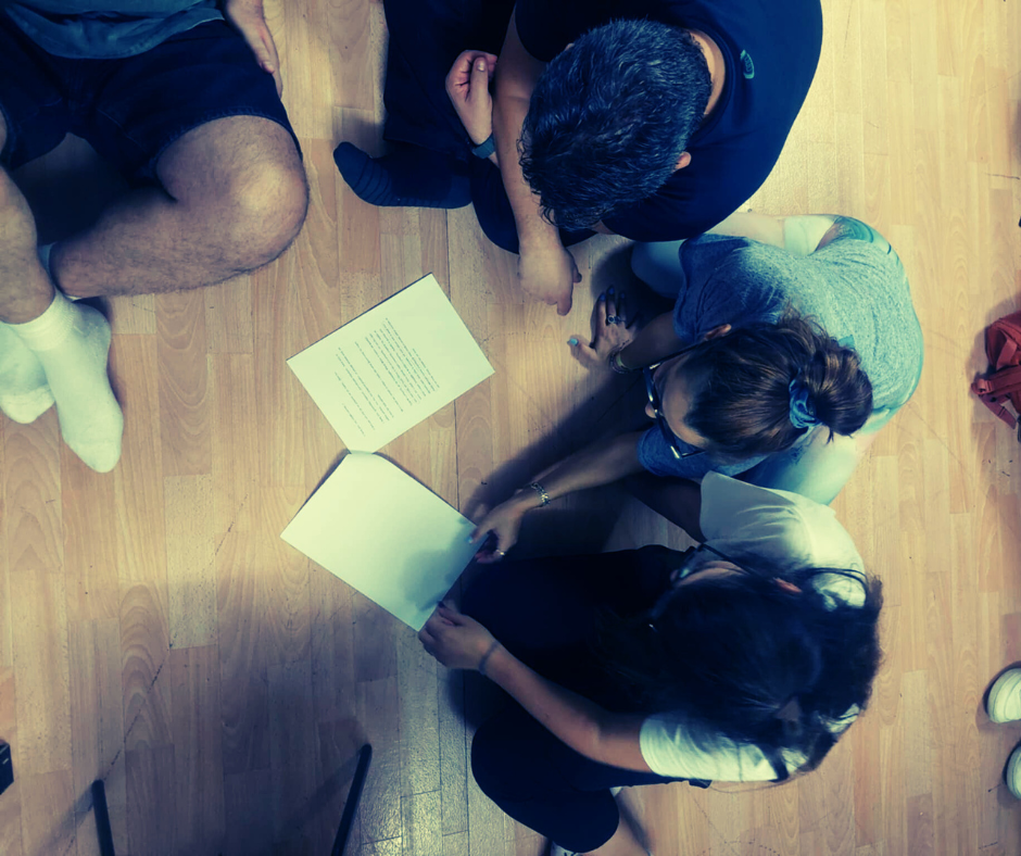 Δημιουργική γραφή site post (1) Τρεις άνθρωποι καθισμένοι στο πάτωμα γύρω από φύλλα χαρτιού με κείμενα, συζητούν και συνεργάζονται πάνω στη γραφή, μέσα σε ζεστή και οικεία ατμόσφαιρα εργαστηρίου δημιουργικής γραφής.