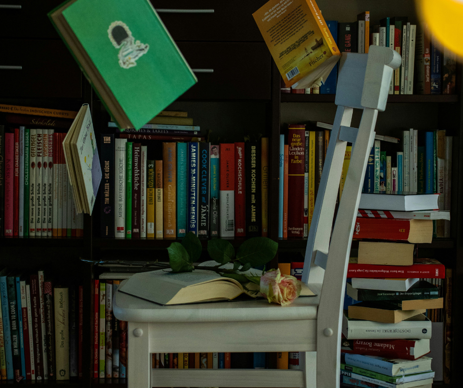 Δημιουργική γραφή site post (2) Μια λευκή καρέκλα μπροστά από βιβλιοθήκη γεμάτη πολύχρωμα βιβλία· πάνω της ένα ανοιχτό βιβλίο και ένα τριαντάφυλλο, ενώ γύρω αιωρούνται βιβλία, δημιουργώντας μια ποιητική ατμόσφαιρα που αποπνέει έμπνευση και αγάπη για τη γραφή.