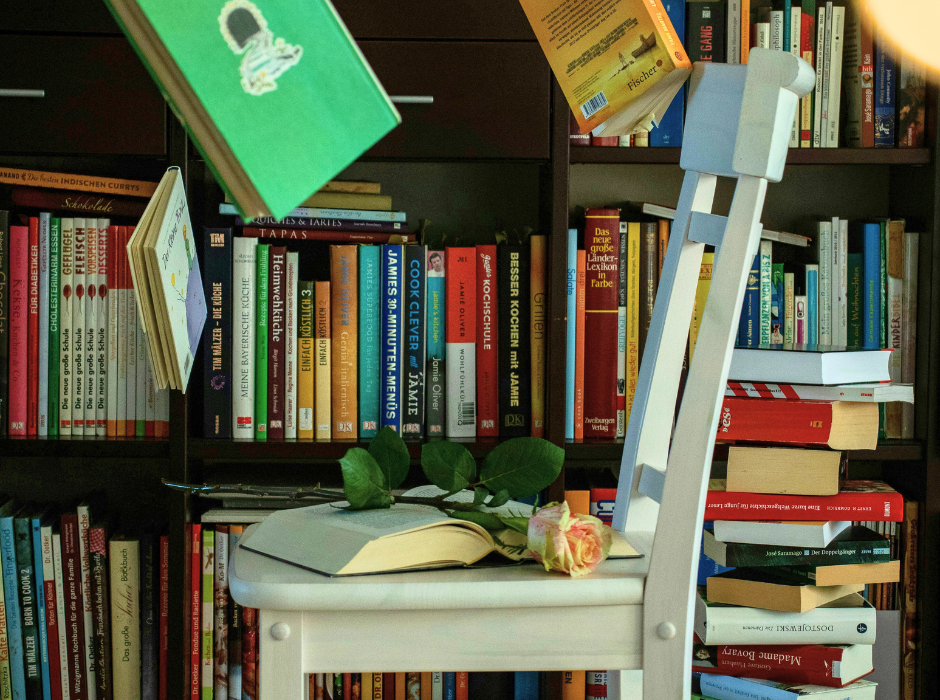 Λευκή ξύλινη καρέκλα τοποθετημένη μπροστά σε μεγάλη βιβλιοθήκη γεμάτη βιβλία. Πάνω στην καρέκλα υπάρχει ανοιχτό βιβλίο με ένα τριαντάφυλλο, ενώ γύρω της αιωρούνται βιβλία, δημιουργώντας μια ονειρική, ποιητική ατμόσφαιρα.