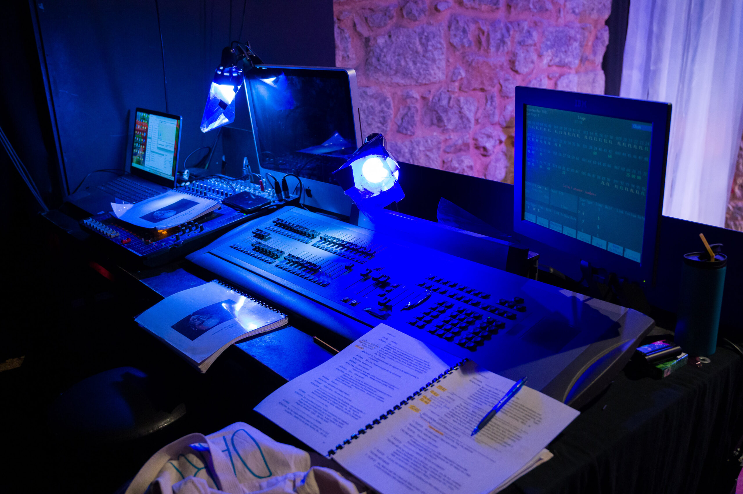 20240628-DSC_0349 Θεατρικό control booth με έντονο μπλε φως. Σε ένα μεγάλο γραφείο βρίσκεται κονσόλα φωτισμού με πολλά faders και κουμπιά, δίπλα σε δύο οθόνες και έναν φορητό υπολογιστή. Πάνω στο τραπέζι υπάρχουν εκτυπωμένες σημειώσεις και κείμενο με μαρκαρισμένα σημεία, καθώς και στυλό. Στο βάθος φαίνεται πέτρινος τοίχος και κουρτίνα.