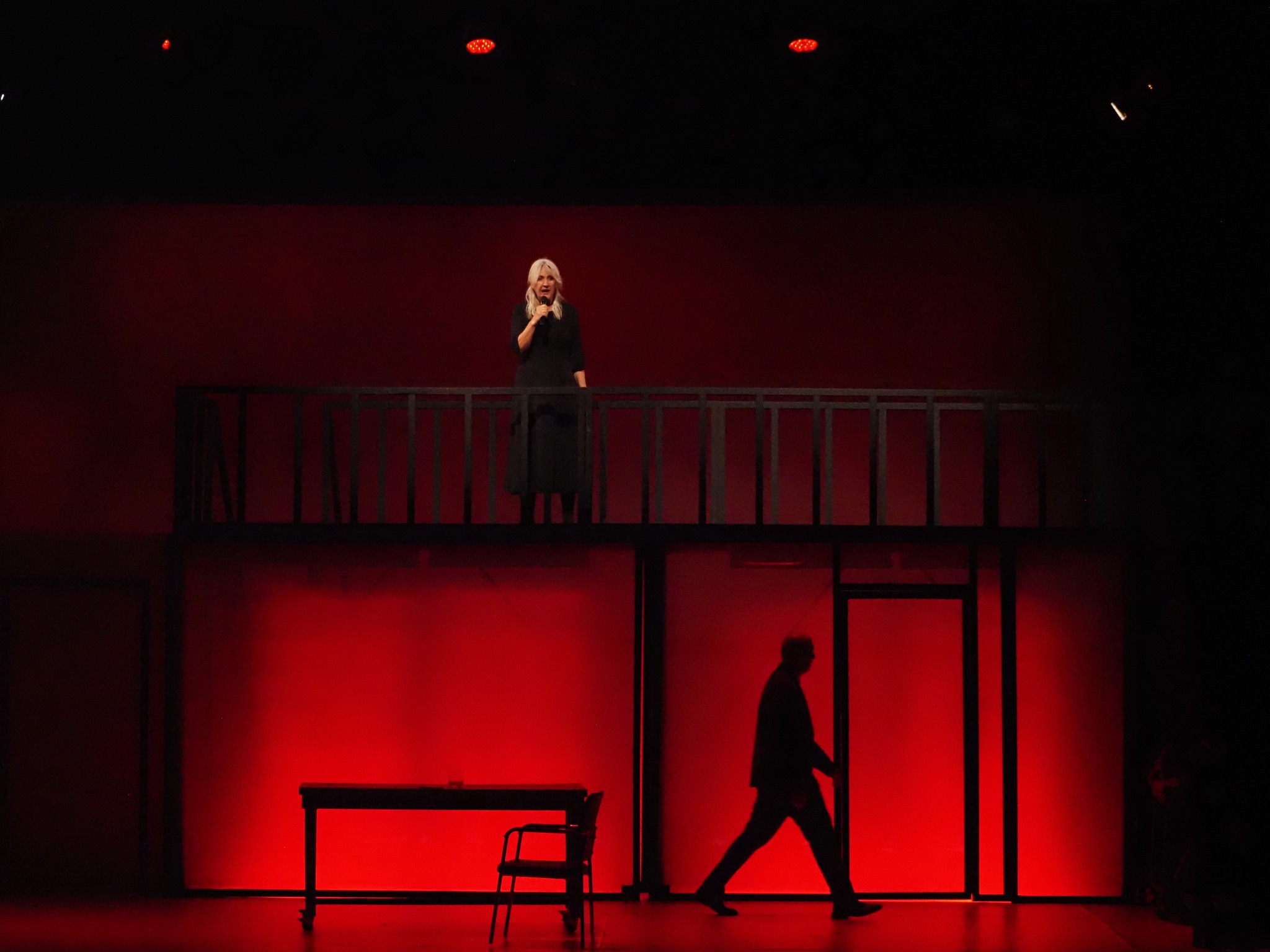 Αμλετ Θεατρική σκηνή φωτισμένη με βαθύ κόκκινο. Στο πάνω επίπεδο, μια γυναίκα με ξανθά μαλλιά και μαύρα ρούχα κρατά μικρόφωνο και κοιτά προς το κοινό πίσω από μαύρο κάγκελο. Στο κάτω επίπεδο φαίνεται ένα τραπέζι και μια καρέκλα, ενώ ένας άντρας περπατά στο βάθος σαν σκοτεινή σιλουέτα, δίπλα σε μια πόρτα.